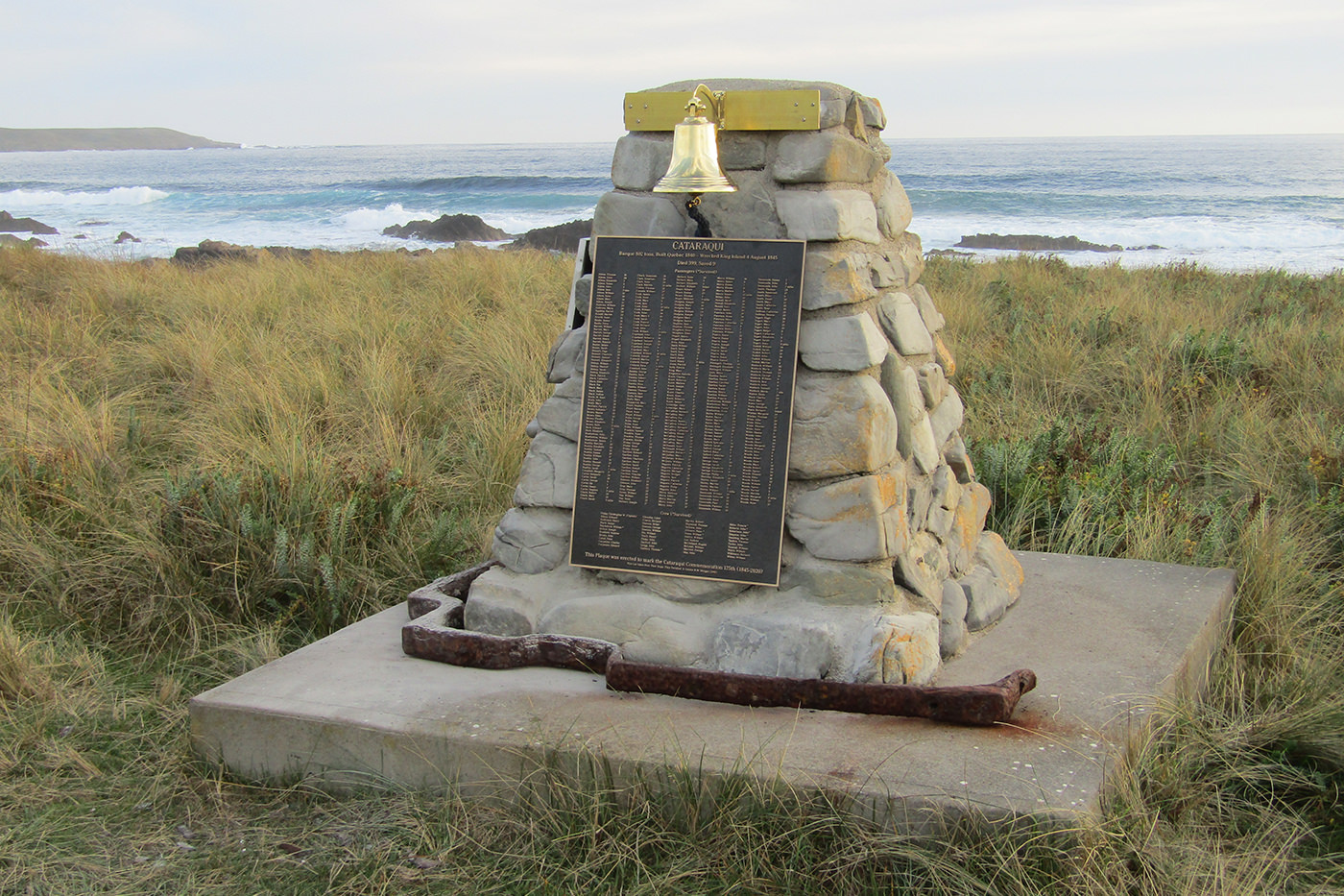 Stone monument with a brass bell and a plaque, surrounded by grass on a rocky shoreline.