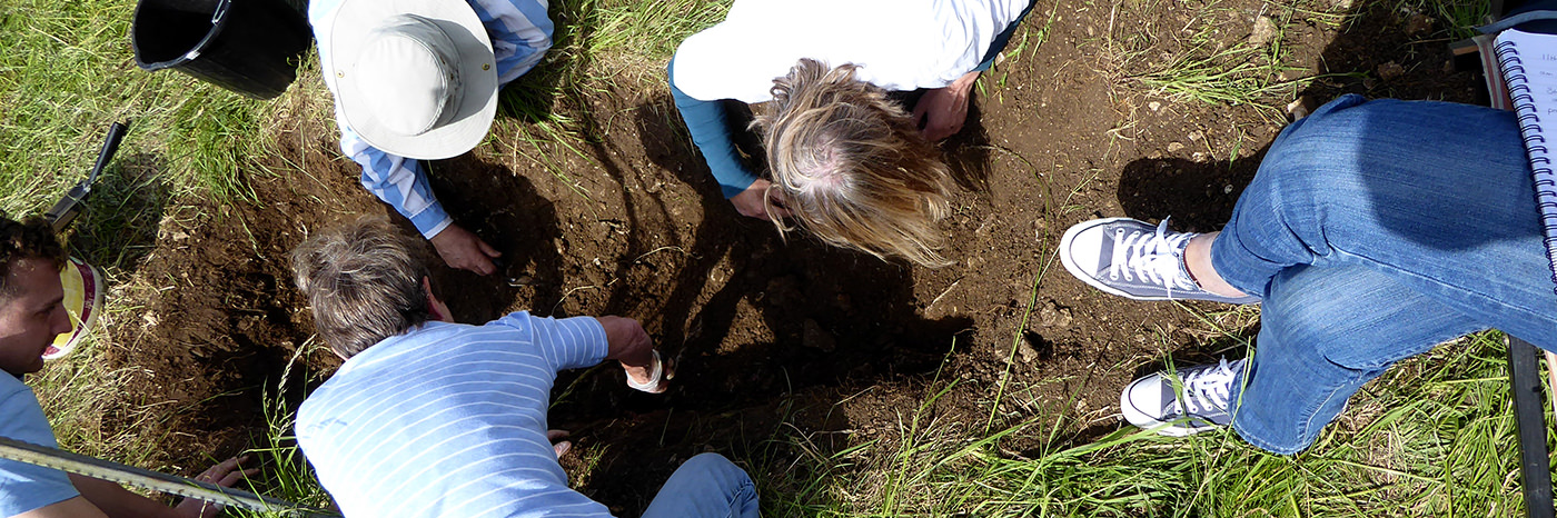 View from above. Three people reach into a pit. Another two look on, one with a notebook.