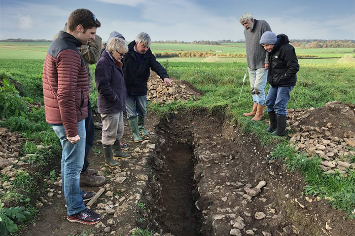 A trench dug in a field, about a metre deep, viewed from one end. Seven people look into it.