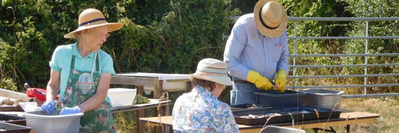 Three people clean artefacts in washing-up bowls. All are wearing hats because it is sunny.