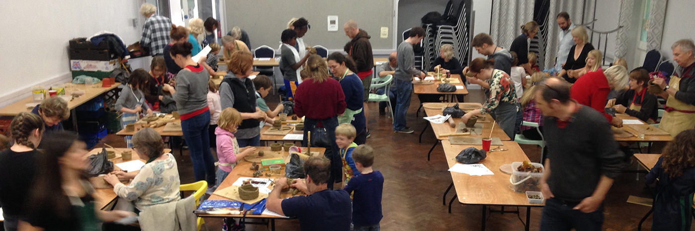 About 30 people of all ages stand around tables working with clay.