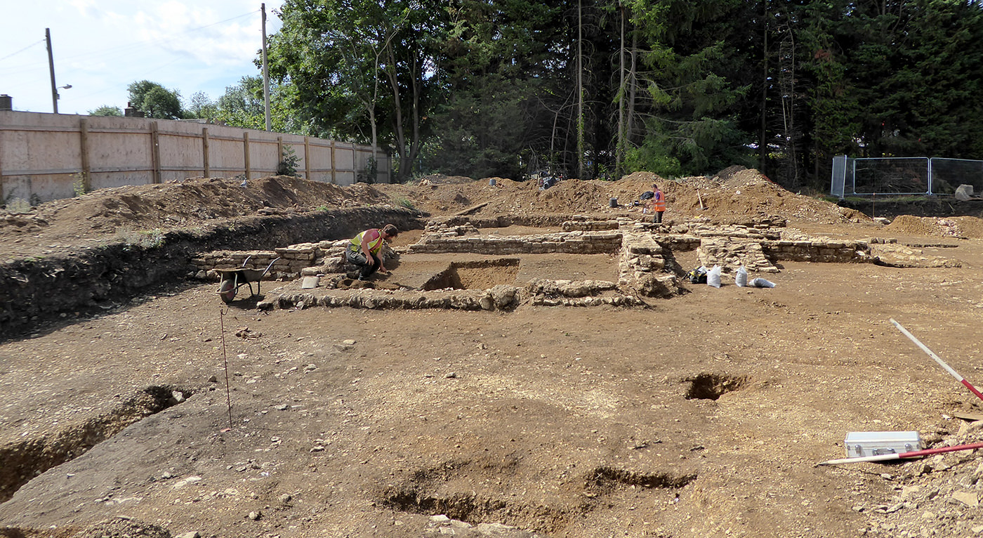 Two archaeologists work at the dig site among pits and unearthed remains.