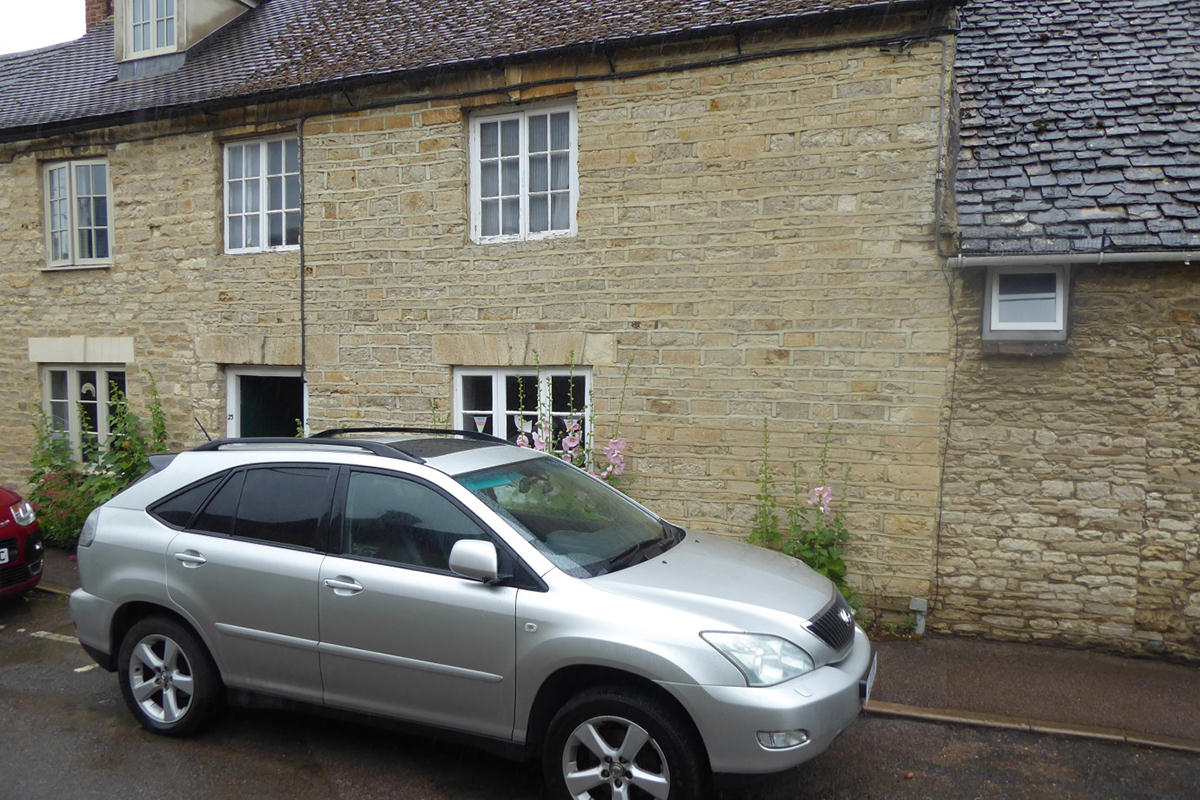 Mid-terrace stone cottage with pink hollyhocks and a silver car outside.