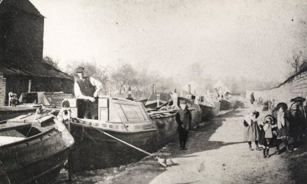 A man stands on board one of several moored canal boats. There are children on the towpath.