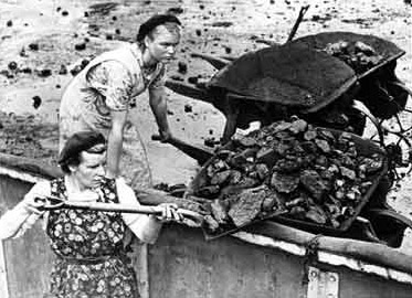 A woman holds the handles of a wheelbarrow while another woman unloads coal from it with a shovel.